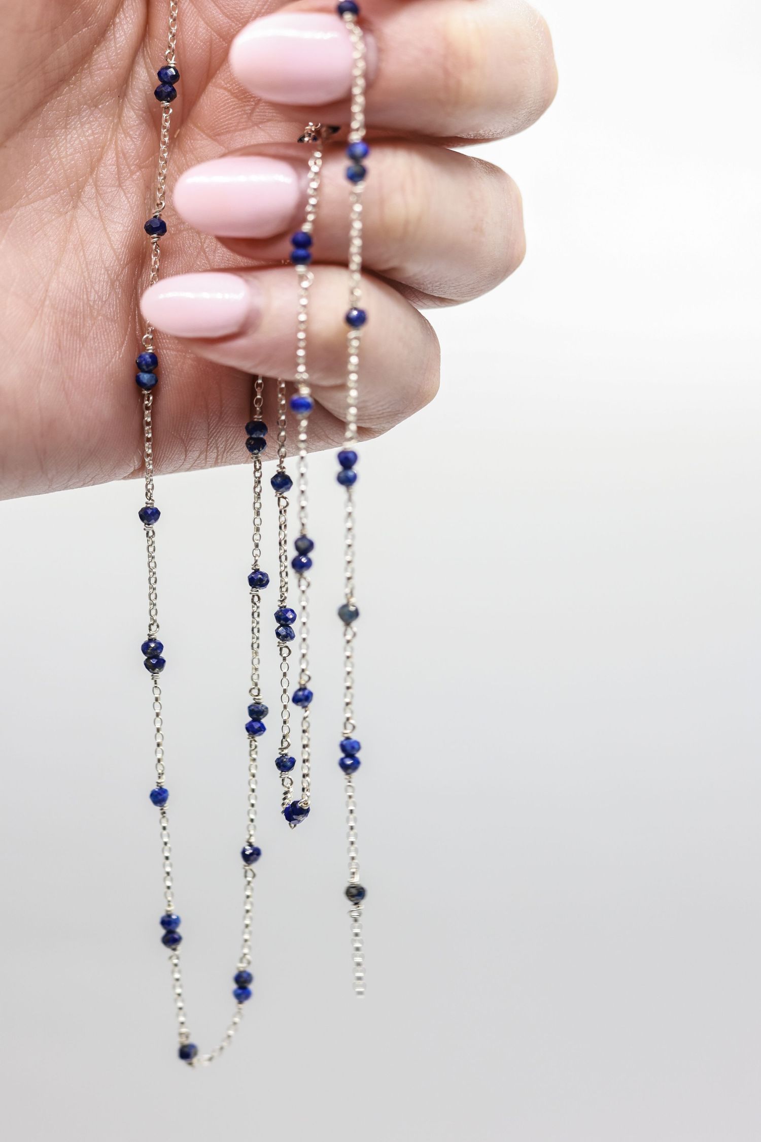 Delicate silver necklace with small blue sapphire beads held by manicured fingers with pale pink nail polish.