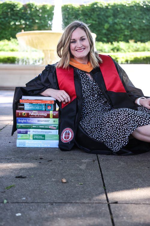 Graduate in academic regalia poses with stack of textbooks near fountain in Abilene, Texas.