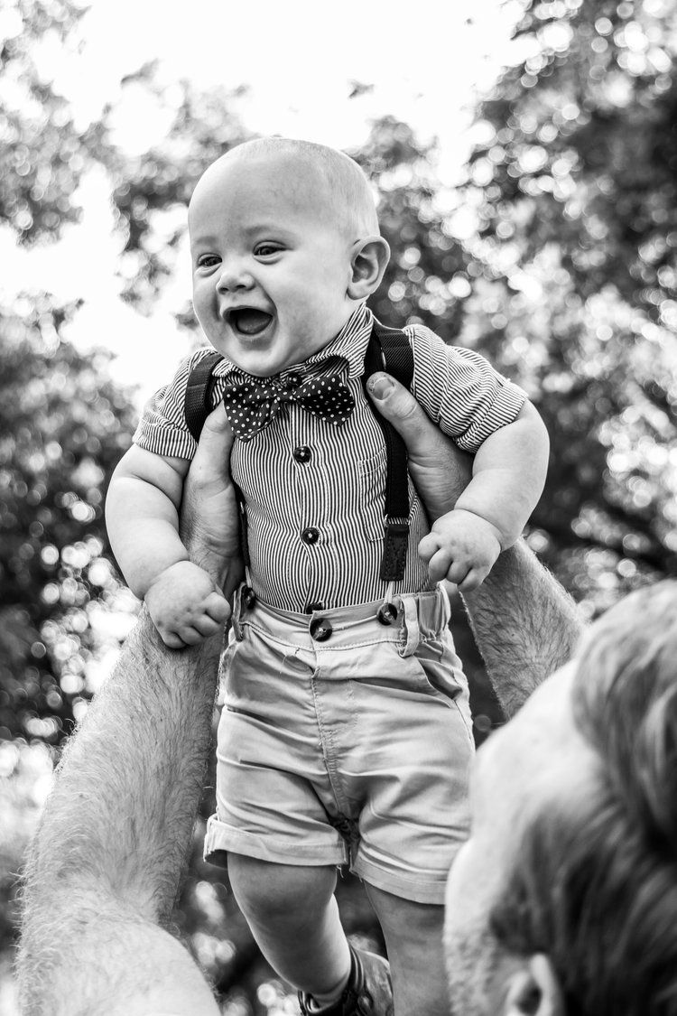 A joyful baby wearing suspenders, bow tie and formal attire is lifted up against a blurred outdoor background in Abilene, Texas.