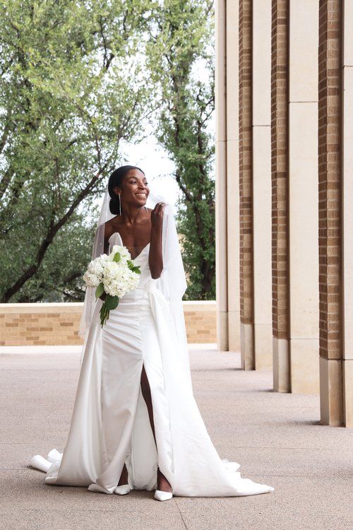 Bride in elegant white gown with flowing cape stands between classic columns  in Abilene, Texas.