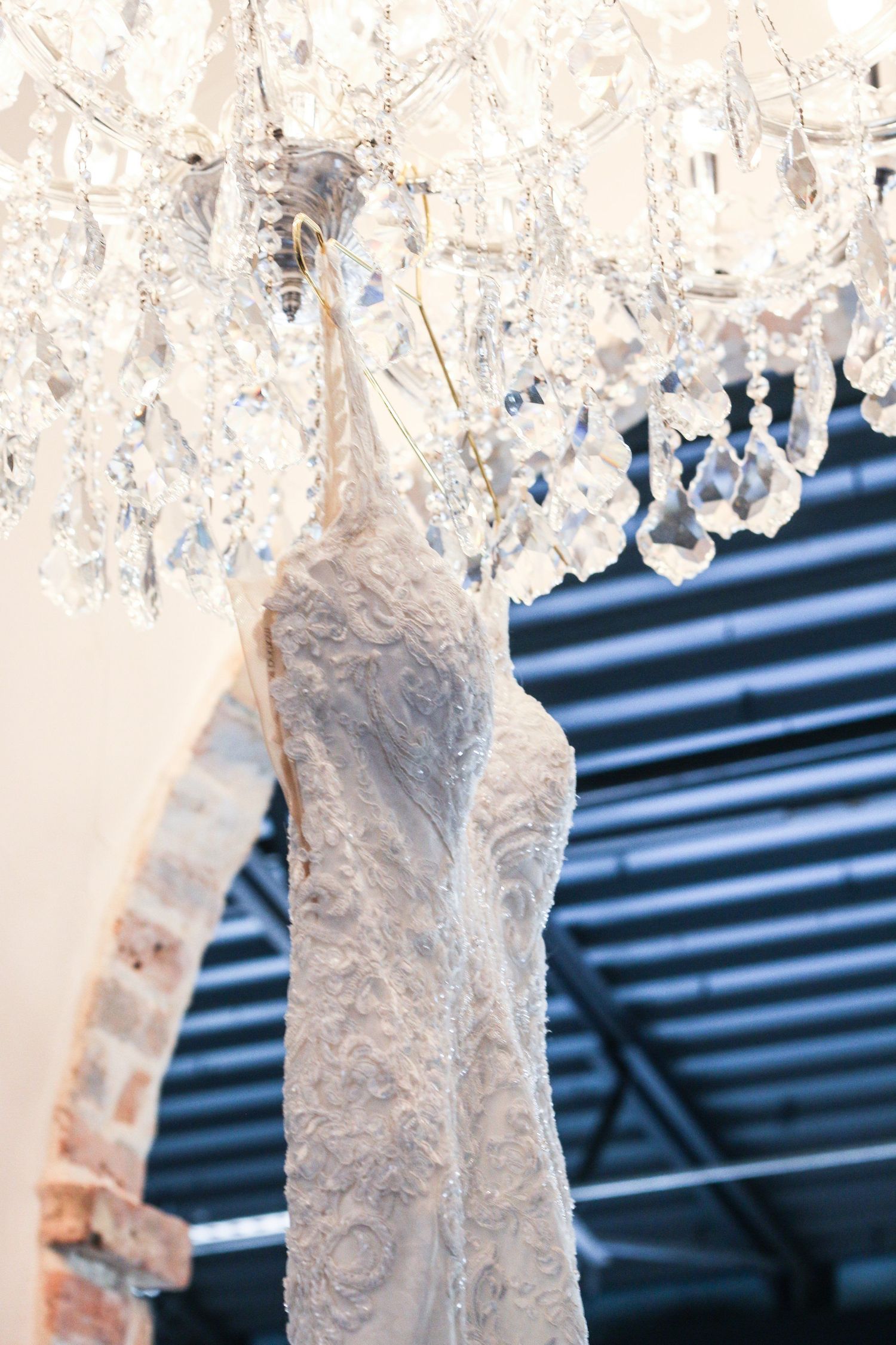 Delicate lace wedding dress hanging from crystal chandelier against dark background.