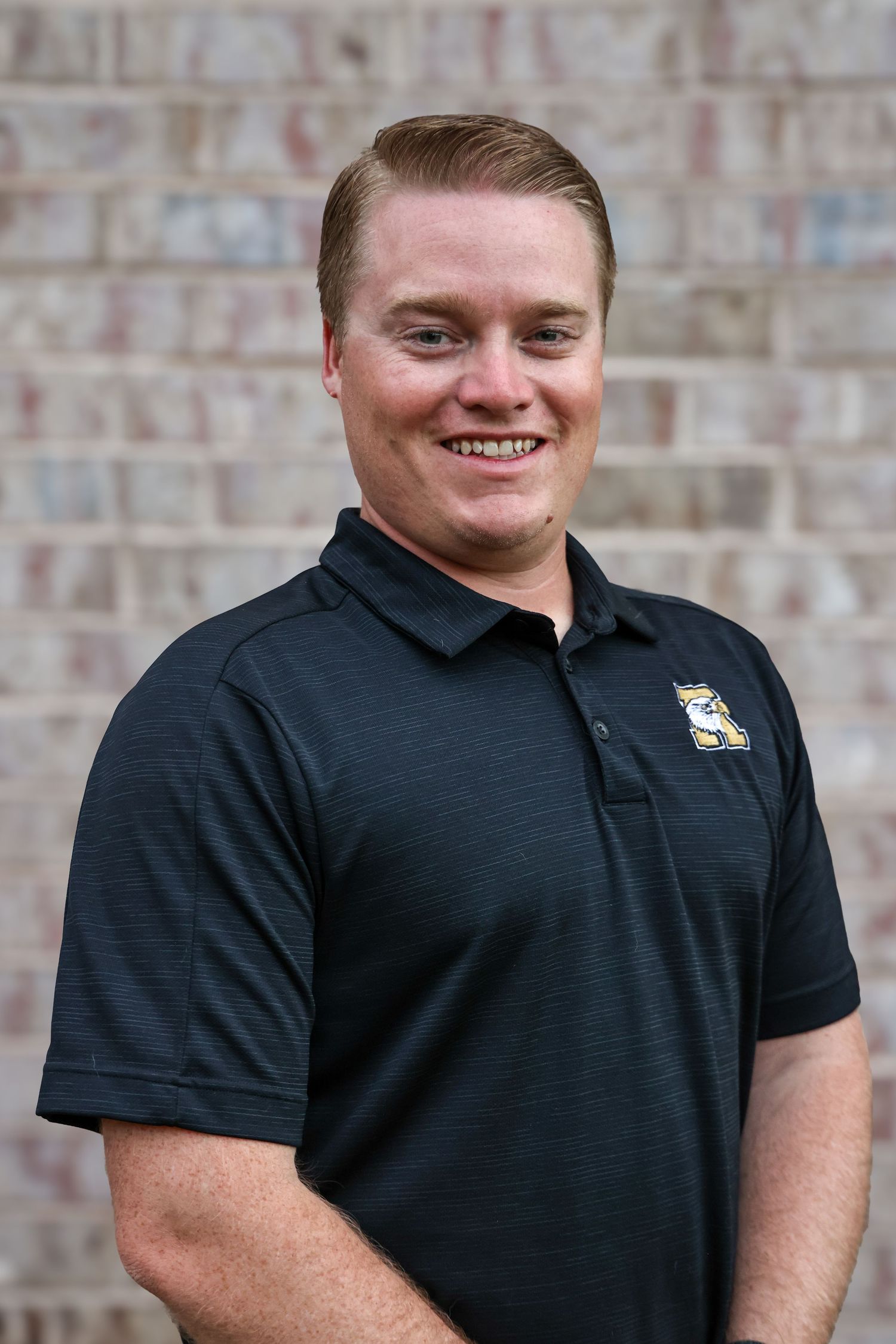 Professional headshot of Abilene coach in a black polo shirt with Eagle logo standing against a brick wall background.