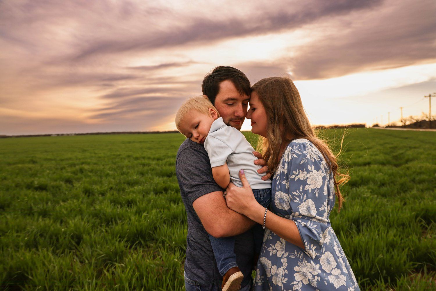 A loving family moment captured at sunset in a green field under dramatic pink clouds in Abilene, Texas.