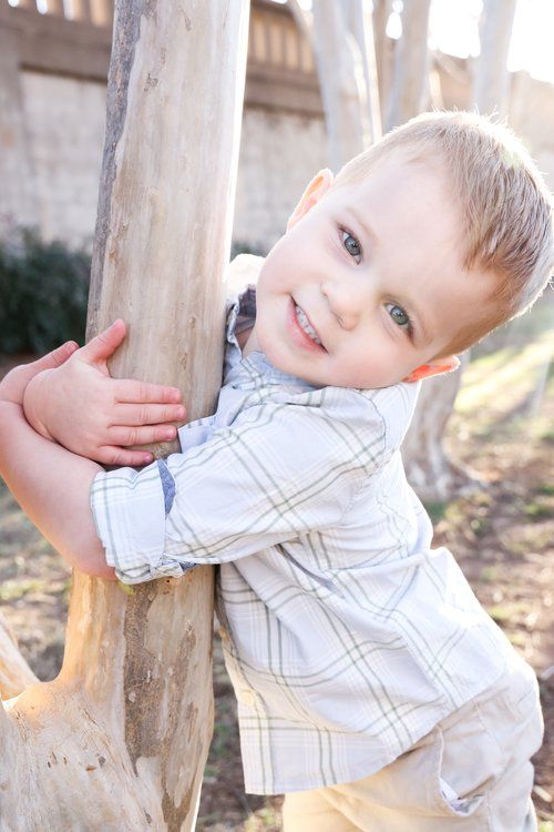 Young child in plaid shirt playfully hugging tree trunk outdoors in Abilene, Texas.