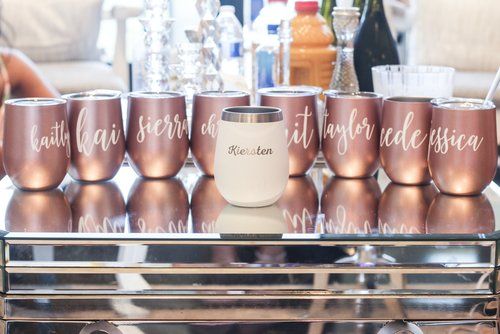 Rose gold wedding party wine cups with custom lettering lined up on a mirrored surface.
