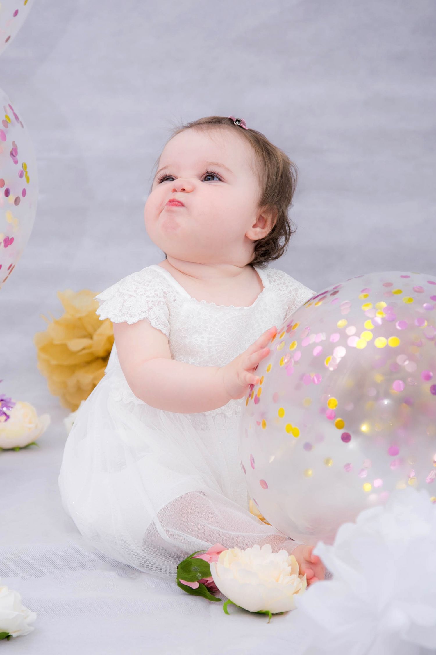 indoor studio cake smash photo shoot of baby in white dress holding a confetti balloon