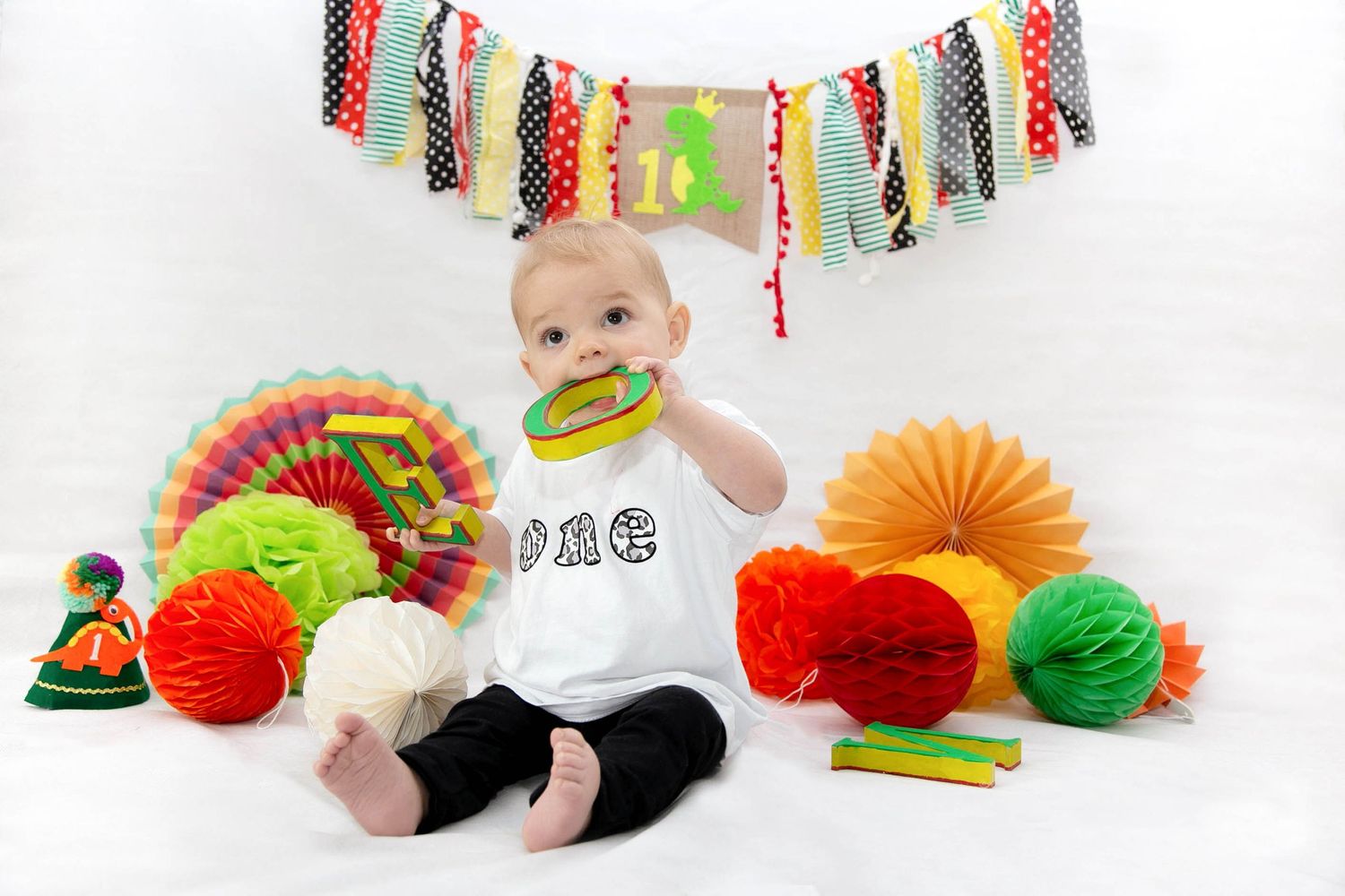 baby chewing on the letters from his first birthday studio photo shoot. brightly coloured props with a dinosaur party hat