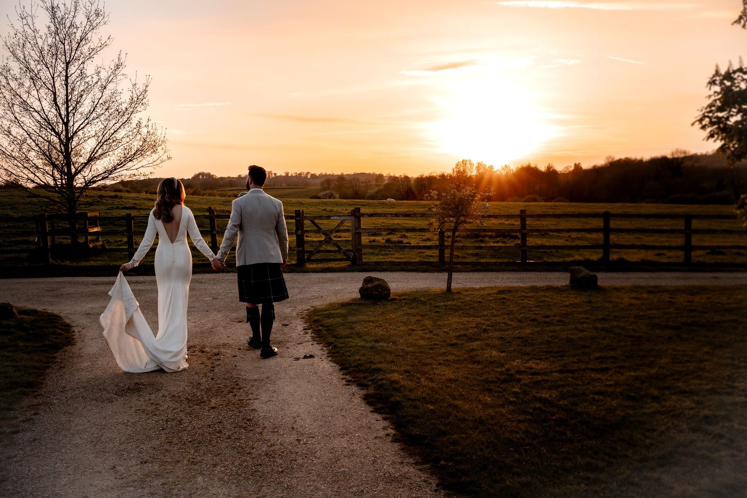Sunset wedding image of bride and groom at dodford manor, walking away into sunset, bride holding her train in one hand. Holding hands