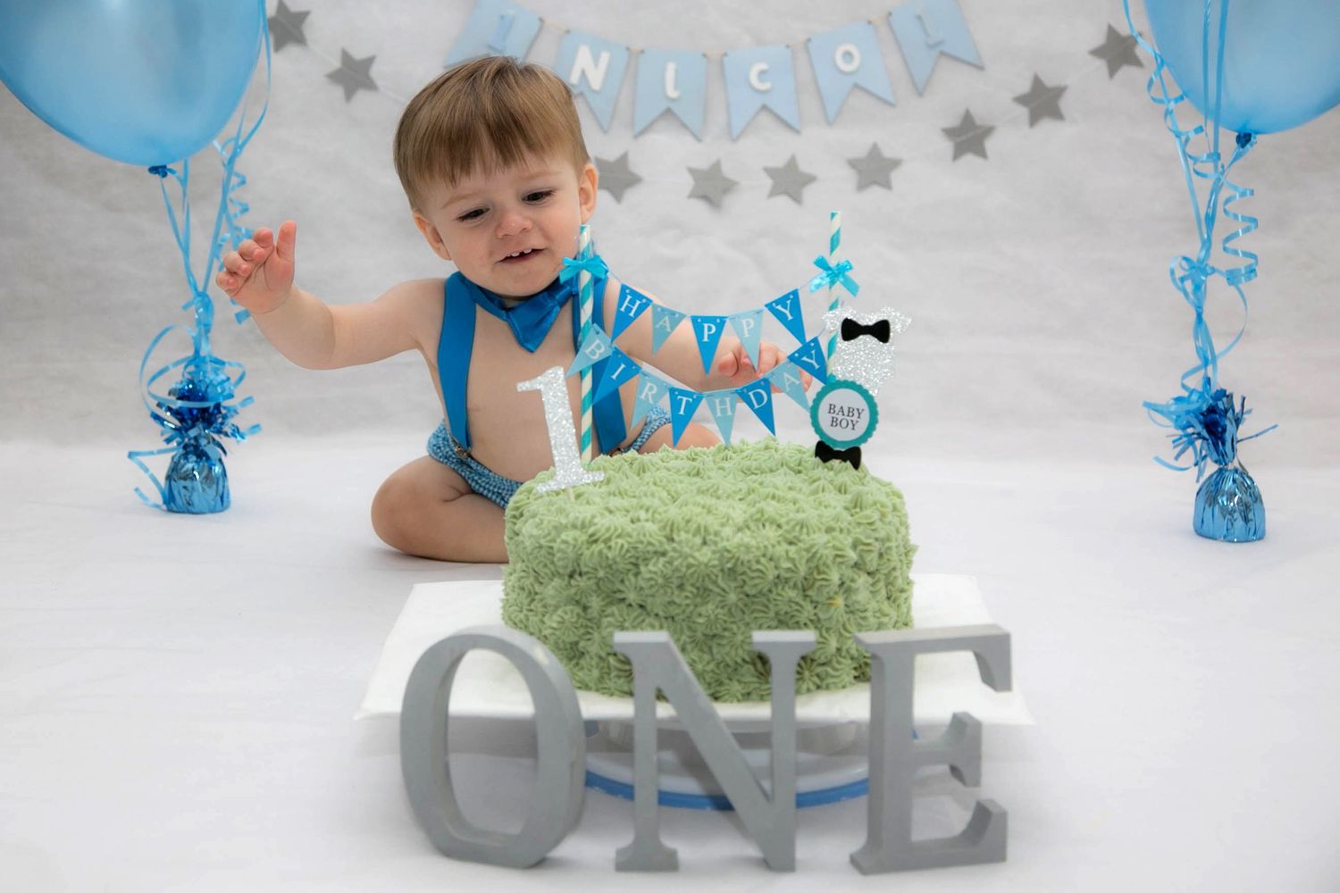 baby boy in blue themed braces for his cake smash photo shoot, with a green cake in front of him with the letters O N E in grey