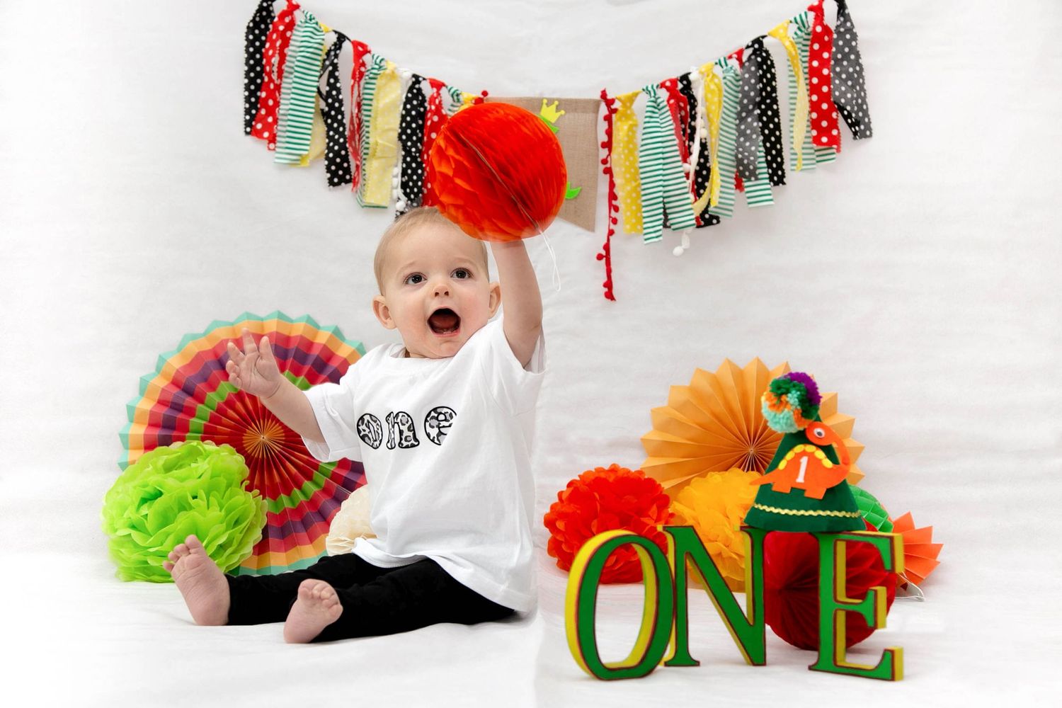 baby boy holding up a red prop in his birthday photo shoot looking happy, sitting next to the wooden letters that spell ONE