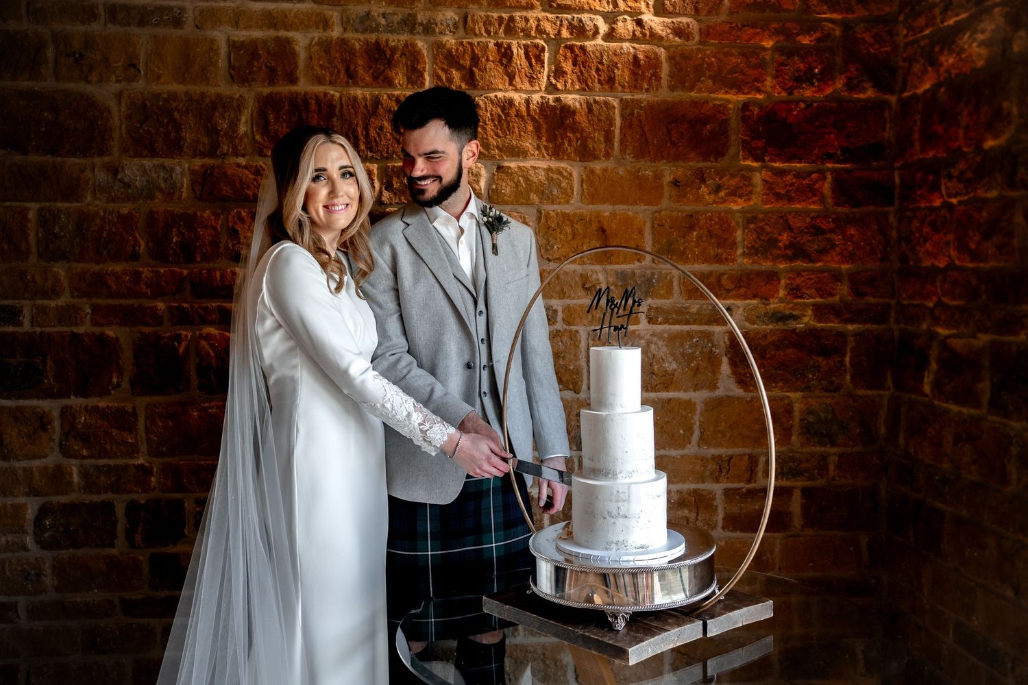 bride and groom cutting their cake at dodford manor. groom smiling and looking at his bride. Brick wall in background