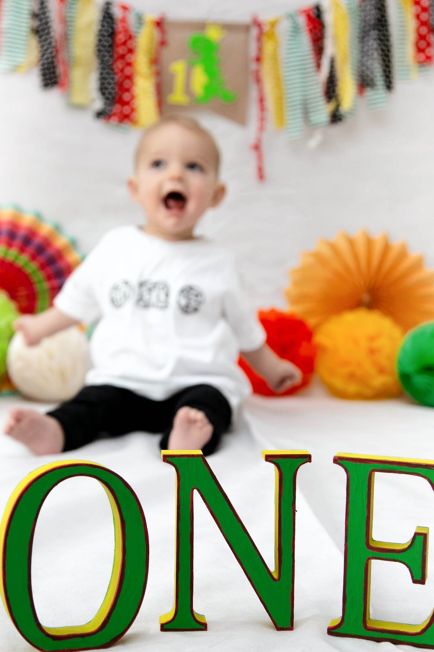 green wooden ONE lettering in foreground of babies first birthday photo shoot. boy in background with large smile on face