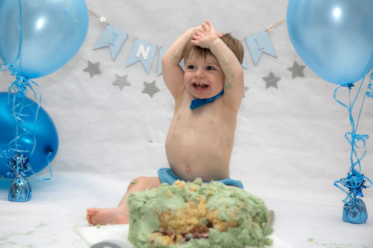 baby smiling and cheering with his hands in the air during his blue themed cake smash session