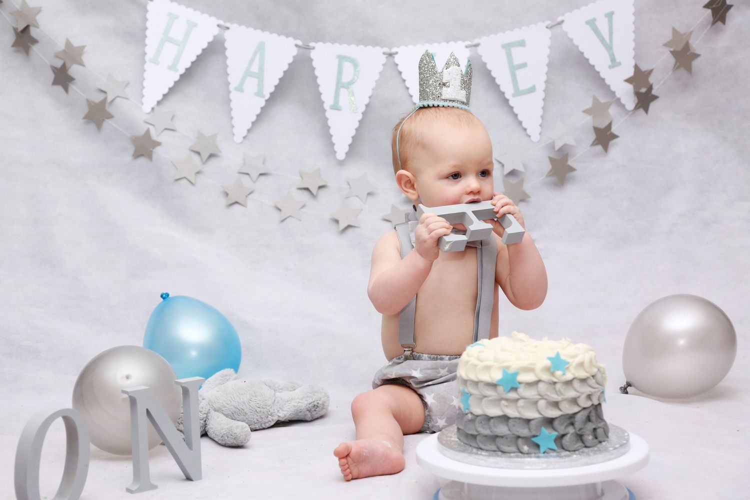 Baby boy in front of grey backdrop with bunting of his name Harley, eating the E wooden lettering prop that spells ONE