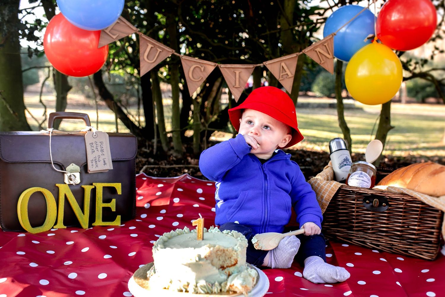 Paddington themed cake smash, one year old boy eating cake and holding a wooden spoon. Balloons and a picnic basket behind him