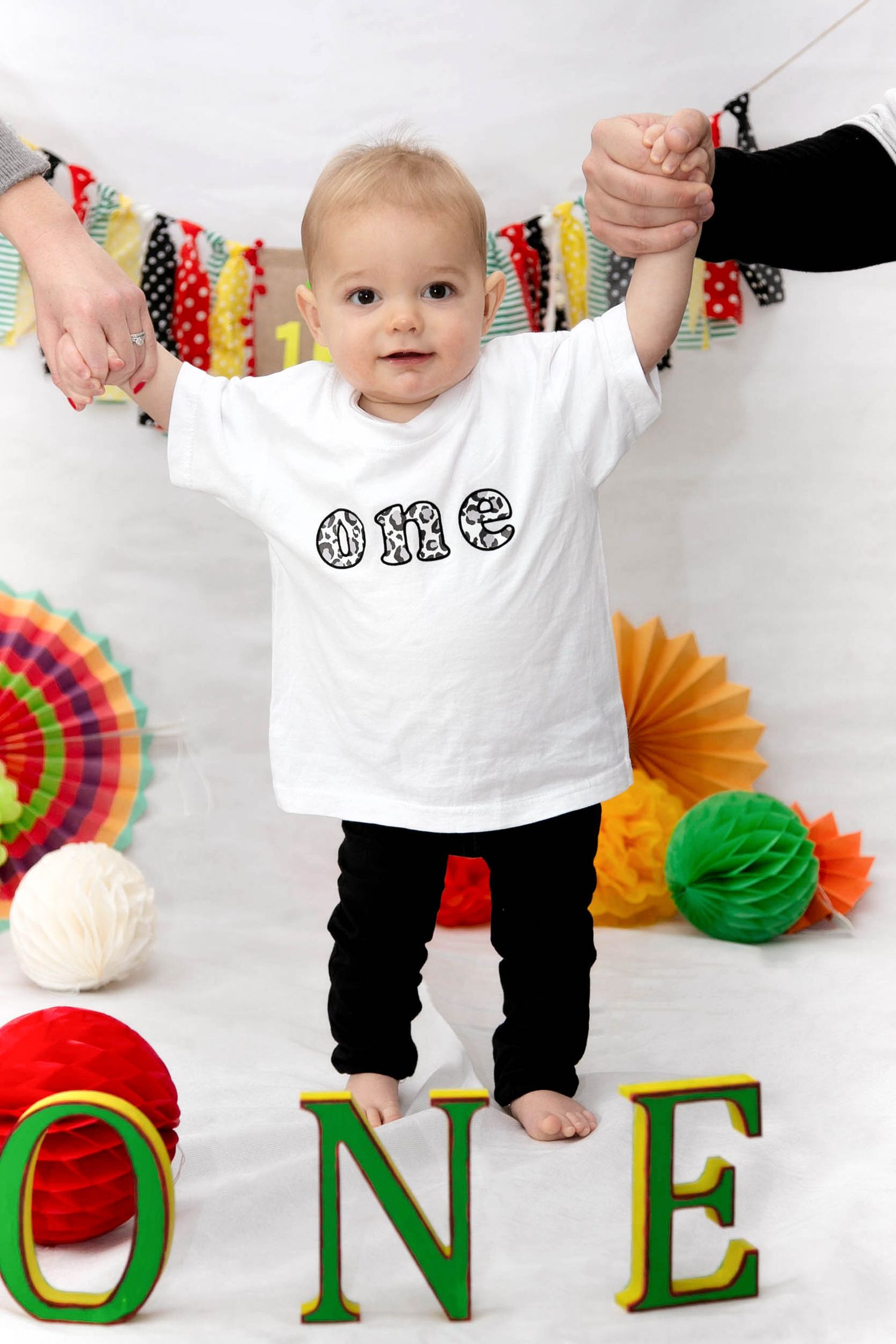 baby standing up being help by mummy and daddy wearing a t-shirt with the words ONE on for his first birthday photo shoot  studio session