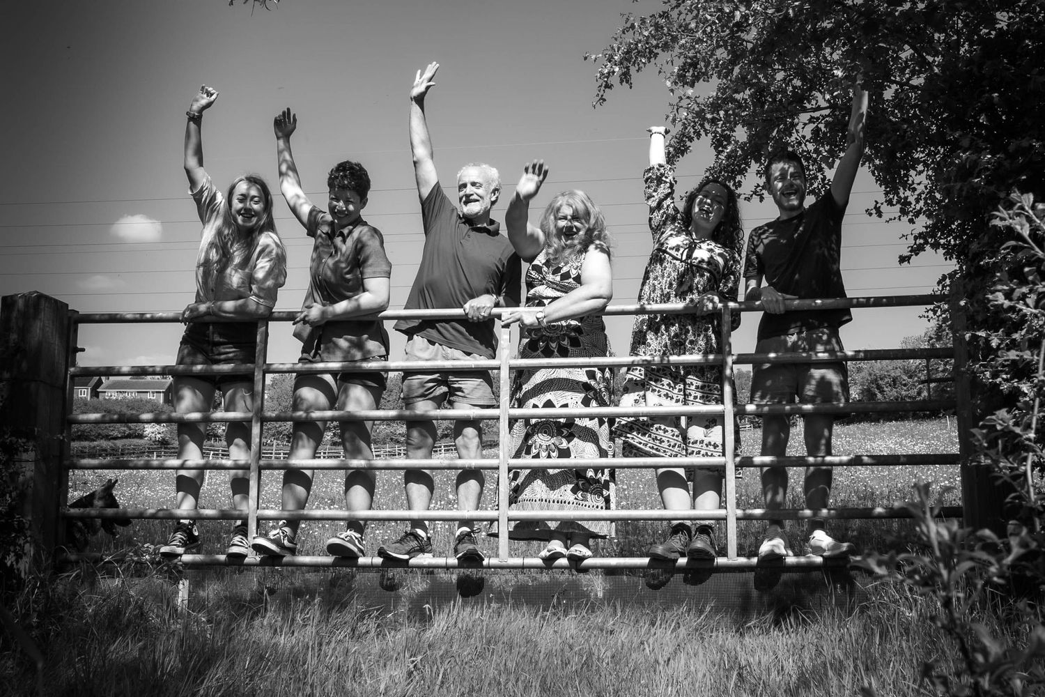 Black and white photo displaying family of six standing on a metal farm gate with their arms in the air smiling and cheering