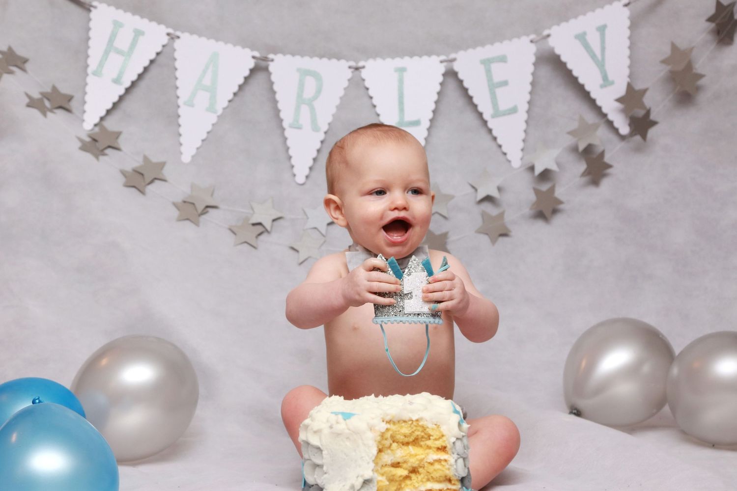 Baby boy in front of grey backdrop with bunting of his name Harley, during his cakesmash session, smiling over to mummy