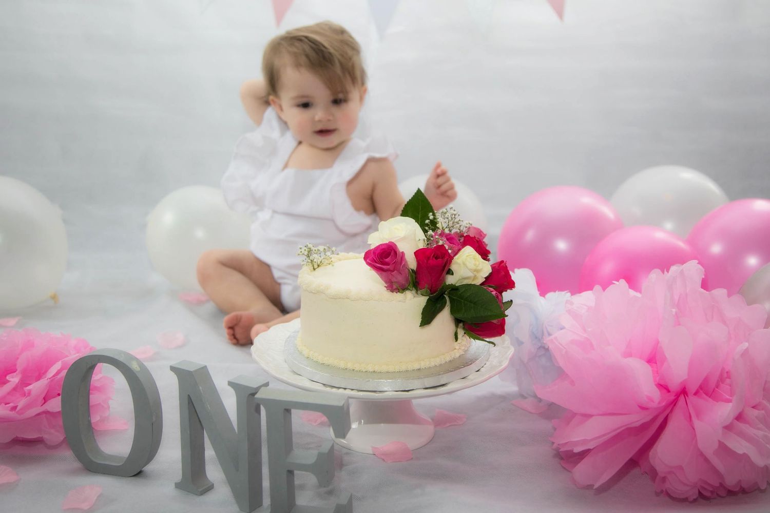 baby cake smash indoor photo shoot. baby looking at cake before eating it. roses on top of cake. pink and white balloons