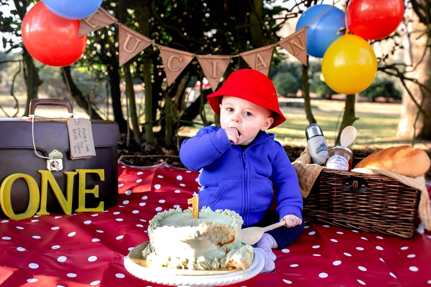 Paddington themed cake smash, one year old boy eating cake and holding a wooden spoon. Balloons and a picnic basket behind him