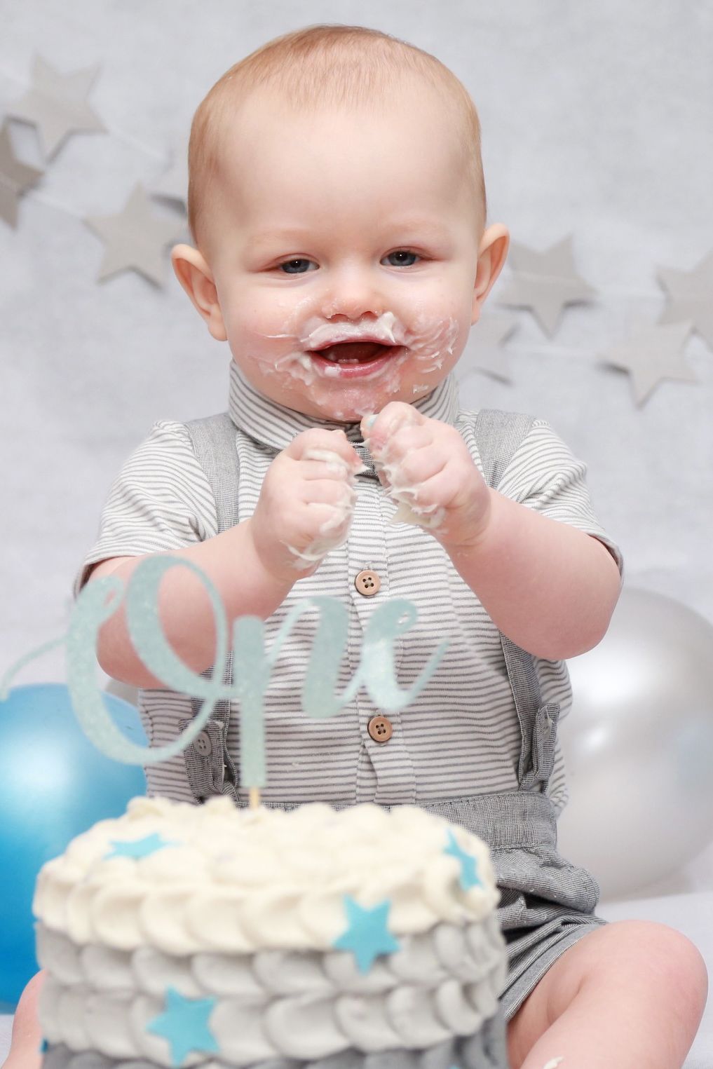 Baby boy eating and enjoying his cake for his cake smash session, wearing grey dungarees