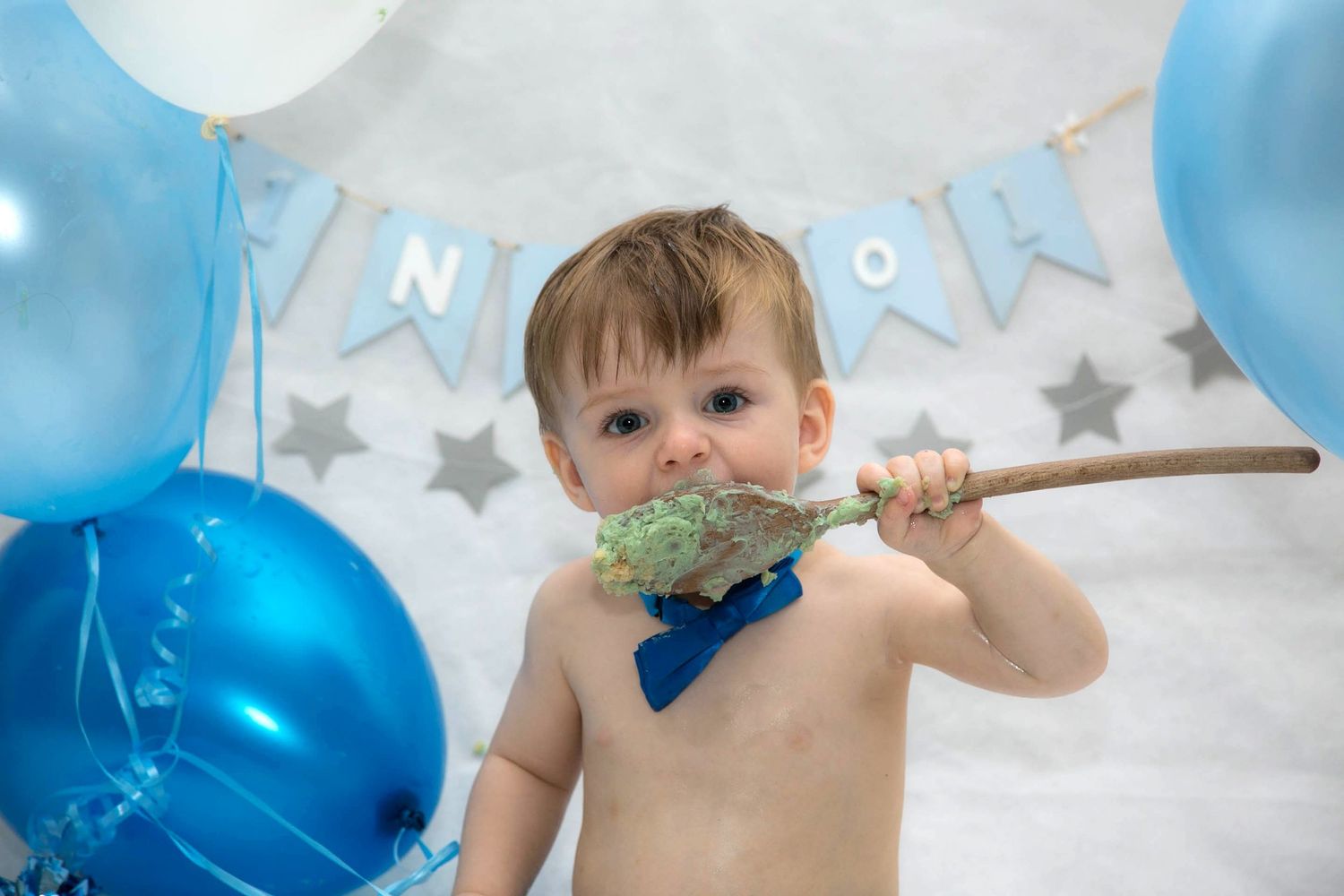 baby boy licking green frosting from a wooden spoon during his blue themed birthday cake smash