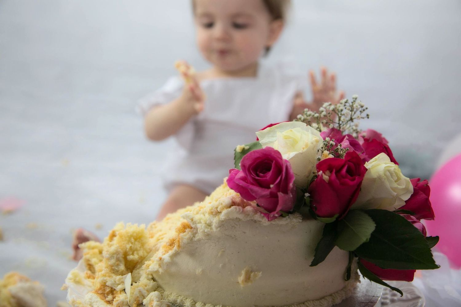 1 year old girl out of focus in background licking icing off her hand, lemon coloured cake in foreground with real pink, white and red roses