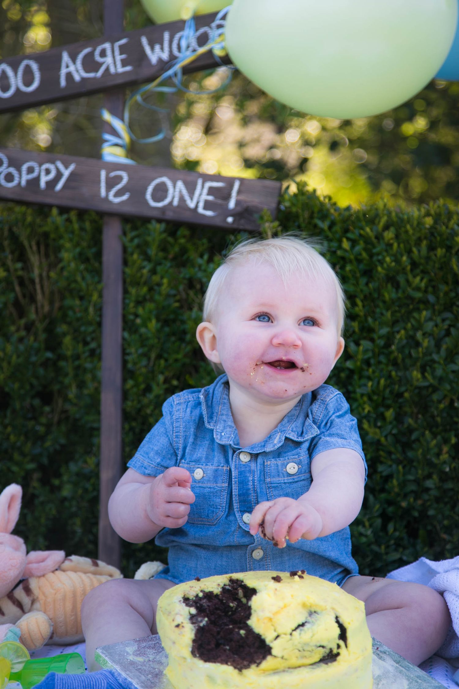 baby girl smiling at mummy and daddy and eating cake outdoors during her winnie the pooh themed cake smash photo shoot