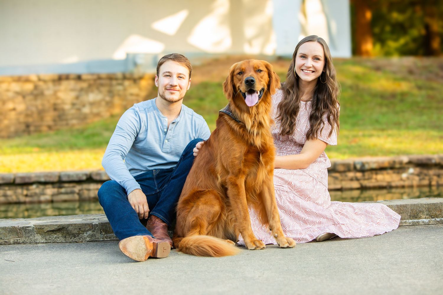 A couple sits with their golden retriever dog on a stone pathway during a sunny outdoor photoshoot.