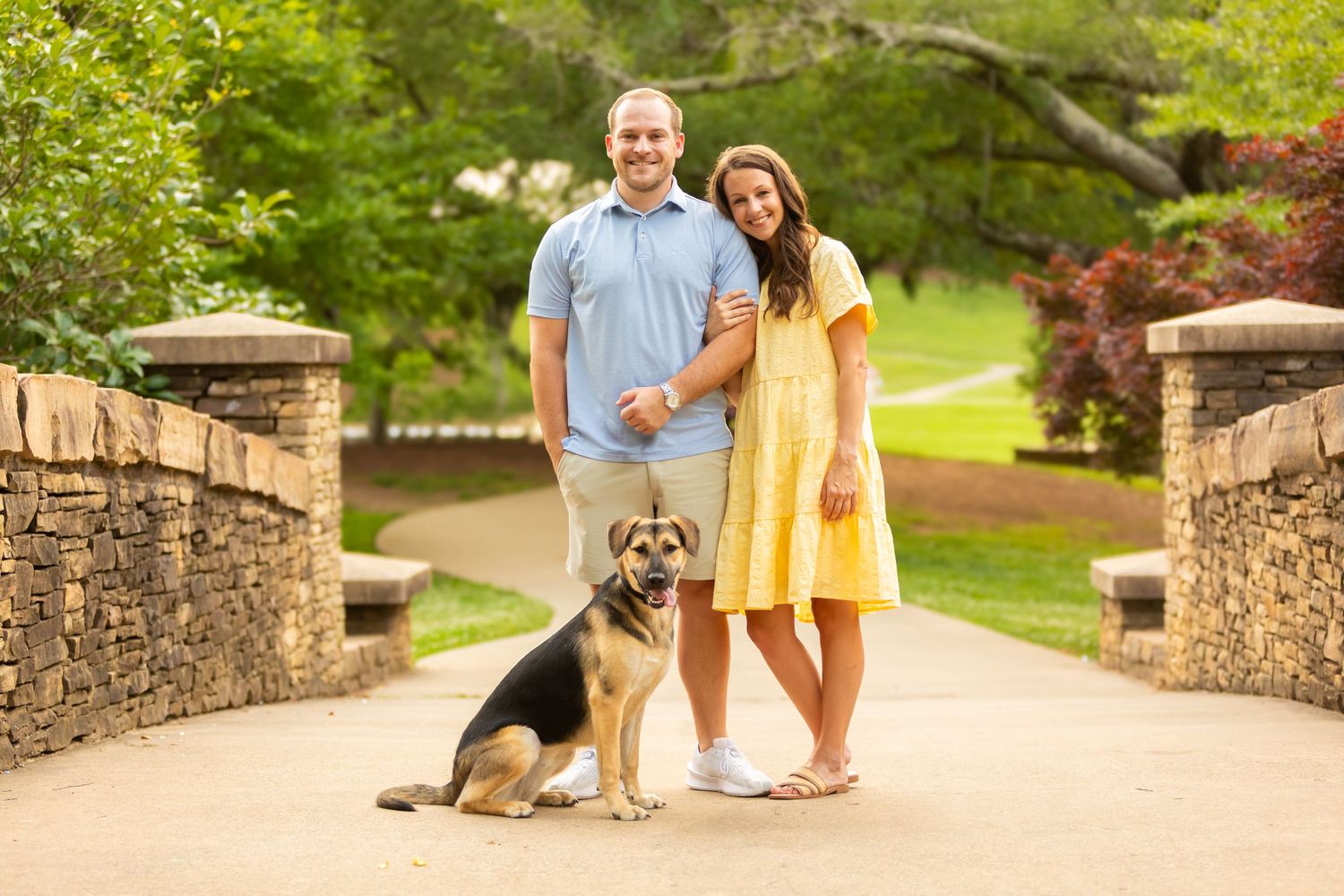 A couple stands on a stone bridge path in a lush garden setting with their large mixed-breed dog.