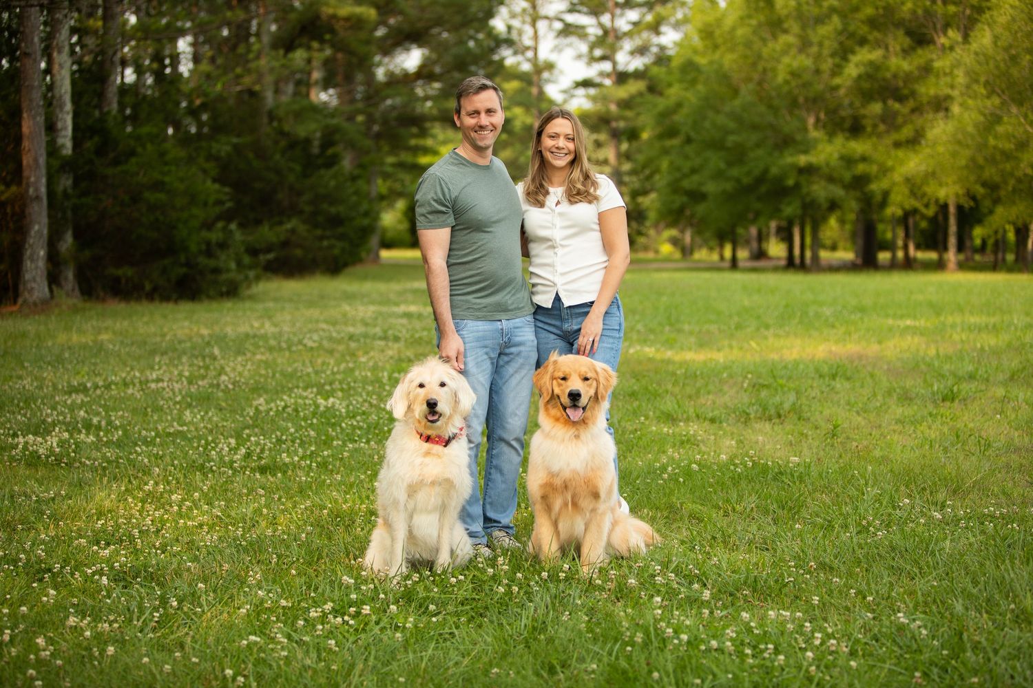 Two Golden Retrievers sitting with their owners on a grassy field with trees in the background.