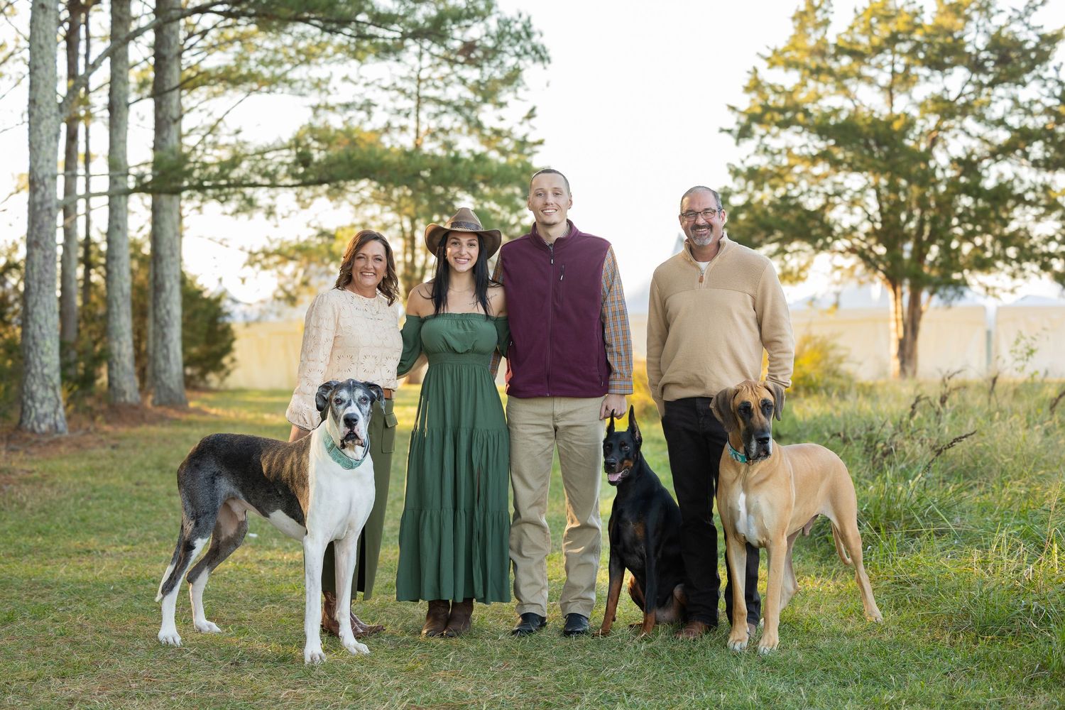 A group of people and their dogs pose together outdoors on a grassy hill with trees in the background at sunset.