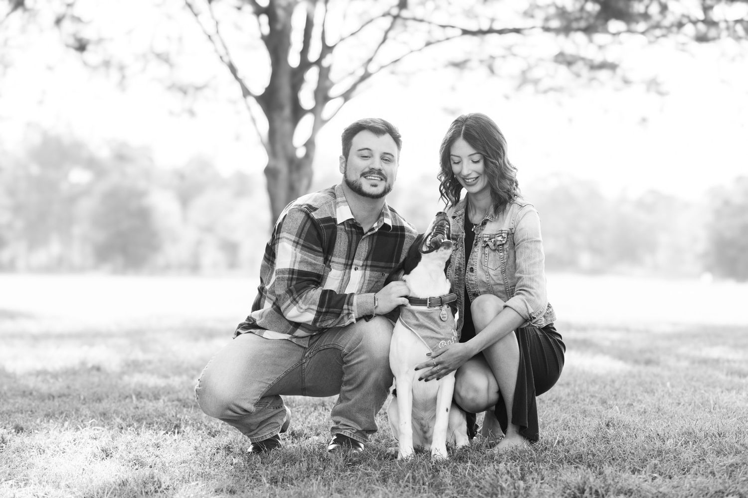 Black and white photo of couple posing with their pet dog in a park setting.