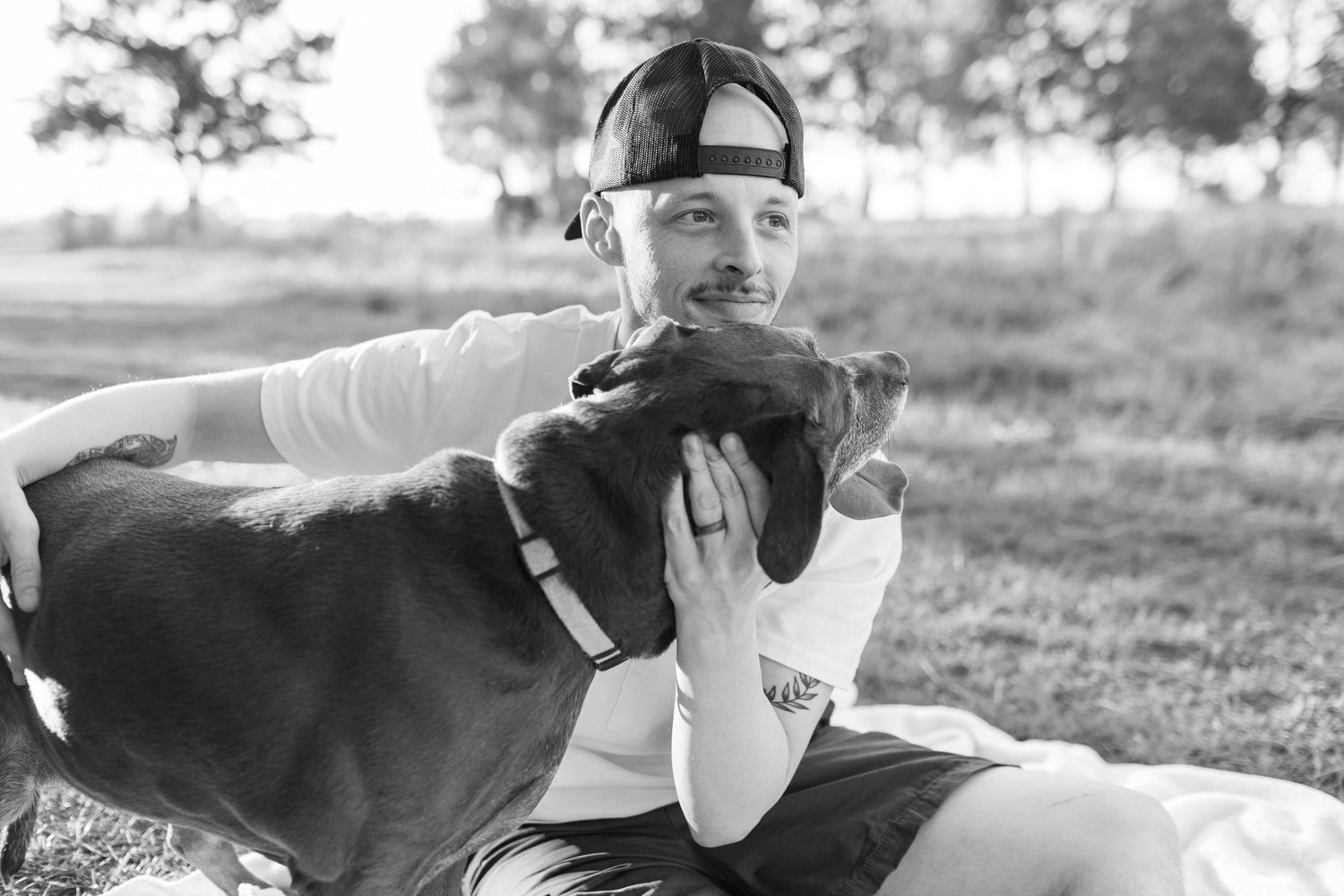 Black and white photo of a young man in a baseball cap affectionately hugging his dog outdoors.