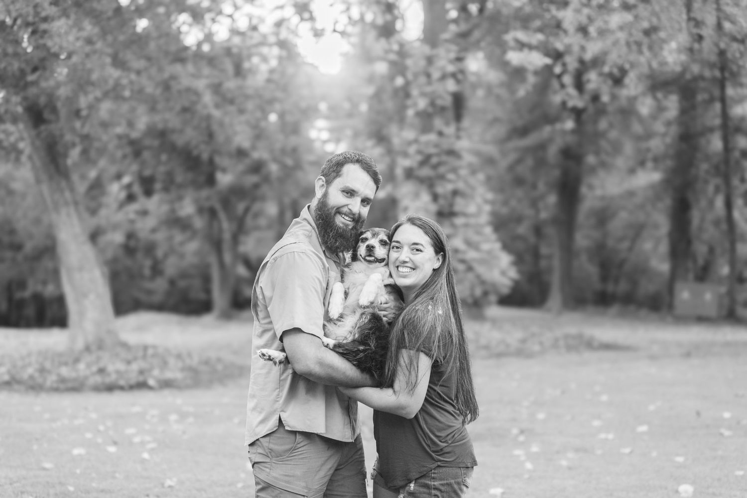 A couple embraces while sharing a tender moment together in a sunlit wooded park setting, captured in black and white.