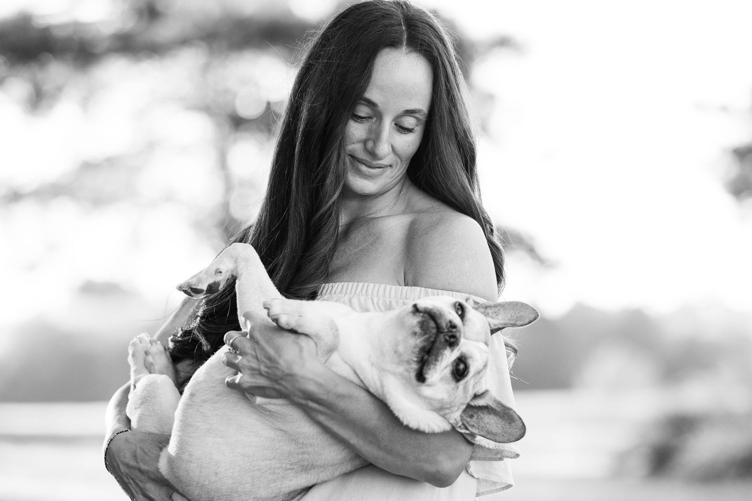 Black and white photo of woman in white dress cuddling French Bulldog while smiling warmly.