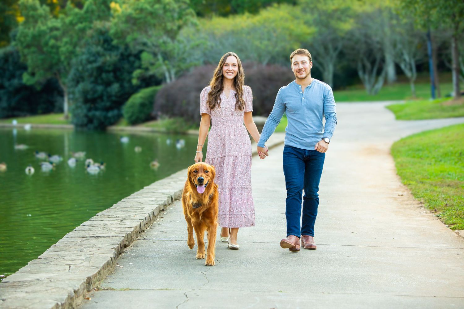 Happy couple walks with their golden retriever dog along a scenic lake path on a sunny afternoon.