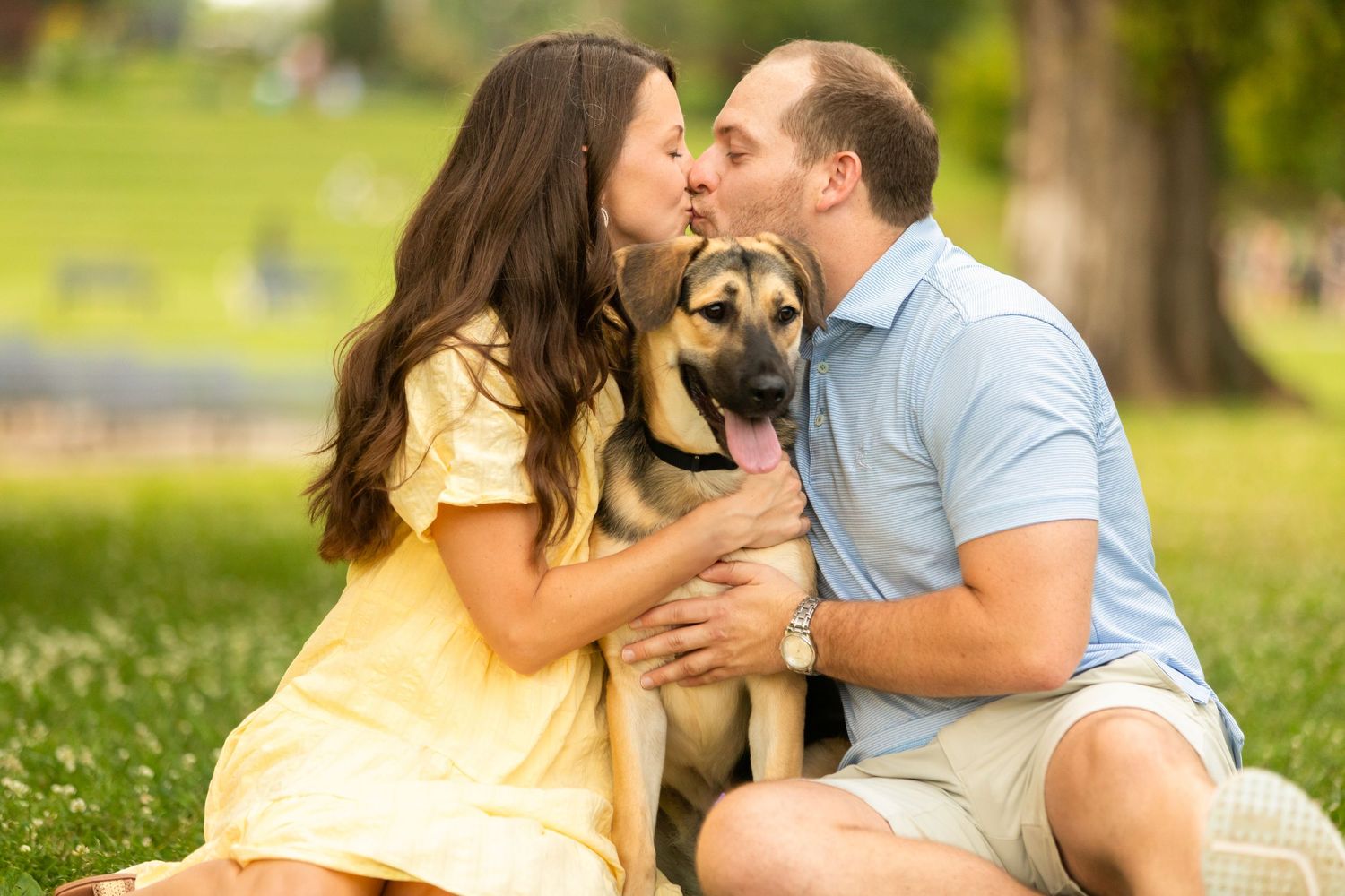 Two people share a tender moment outdoors in a park while holding their German Shepherd mix dog.