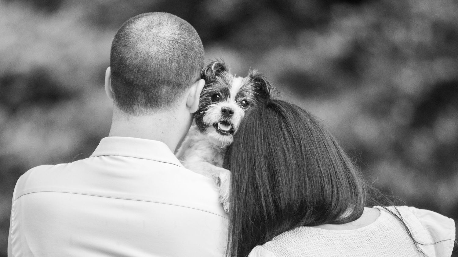 A small white and brown dog looks over a couple's shoulders during an intimate black and white outdoor moment.