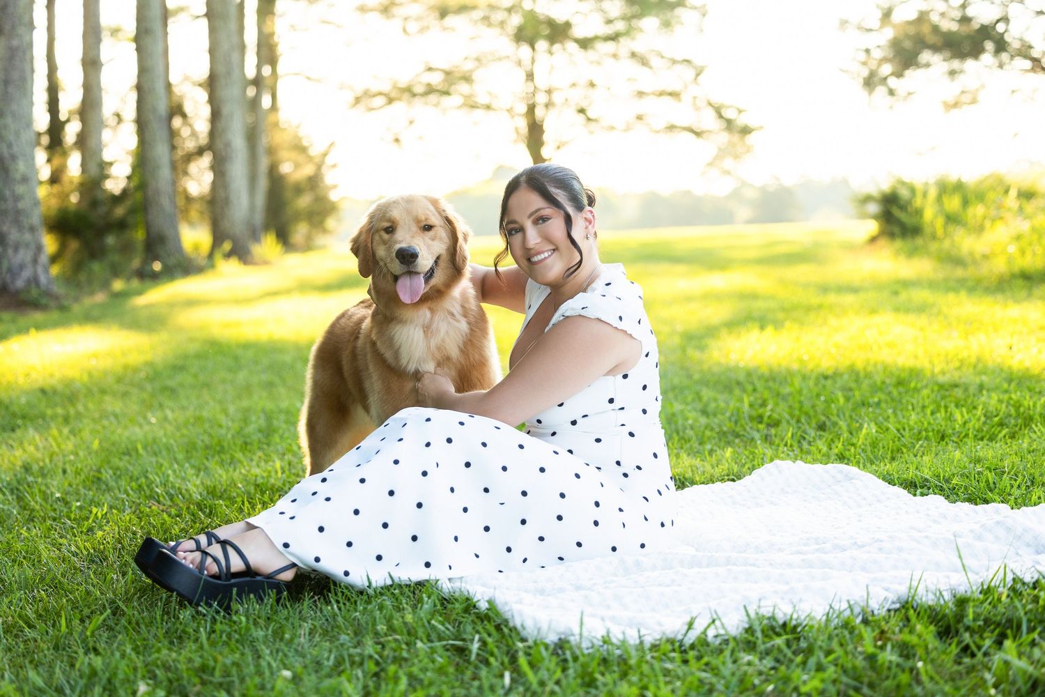 Woman in polka dot dress sitting on grass with golden retriever dog in sunny park setting.