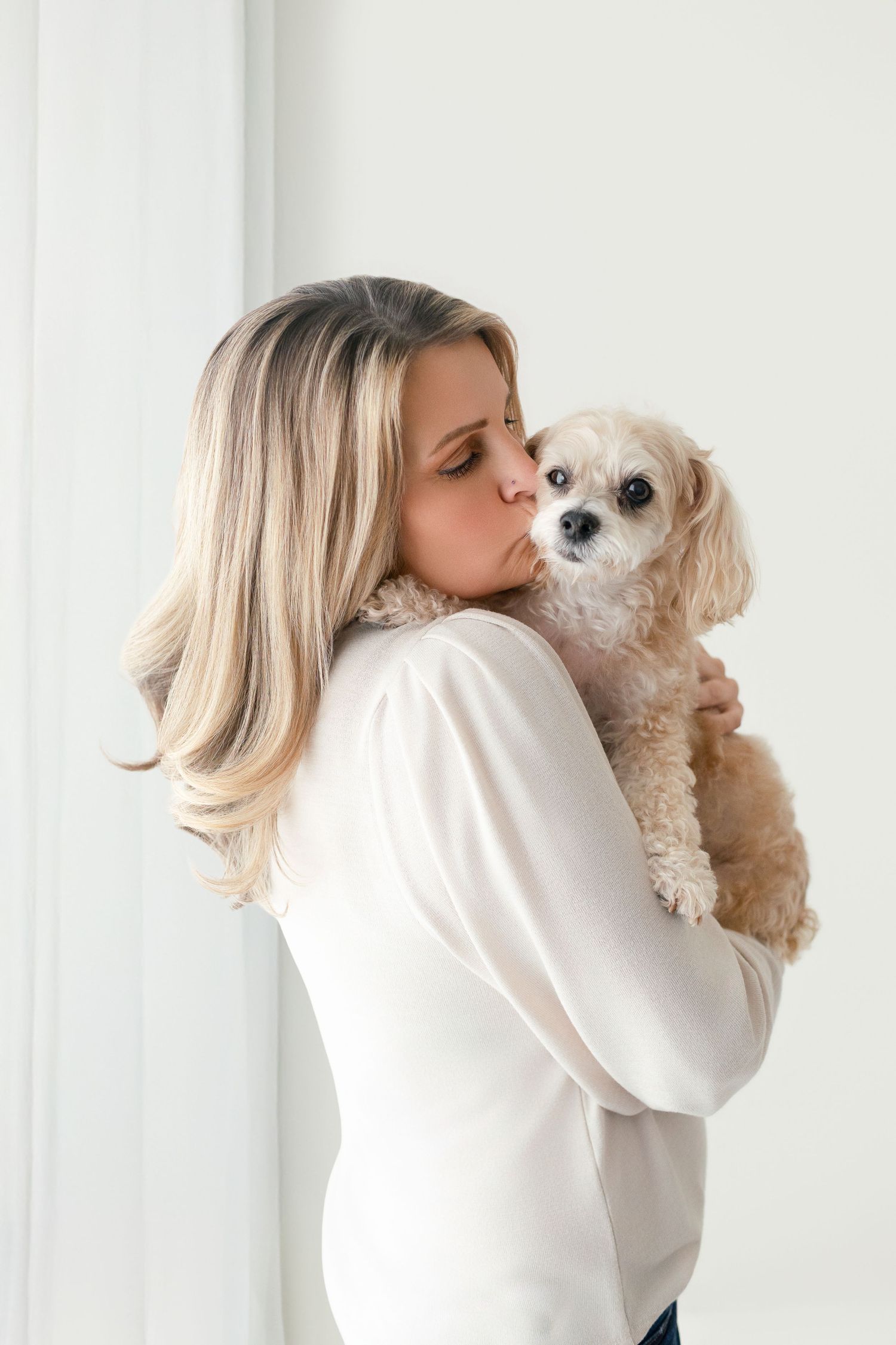 Woman in white sweater kissing her small fluffy Shih Tzu dog while holding it in her arms.