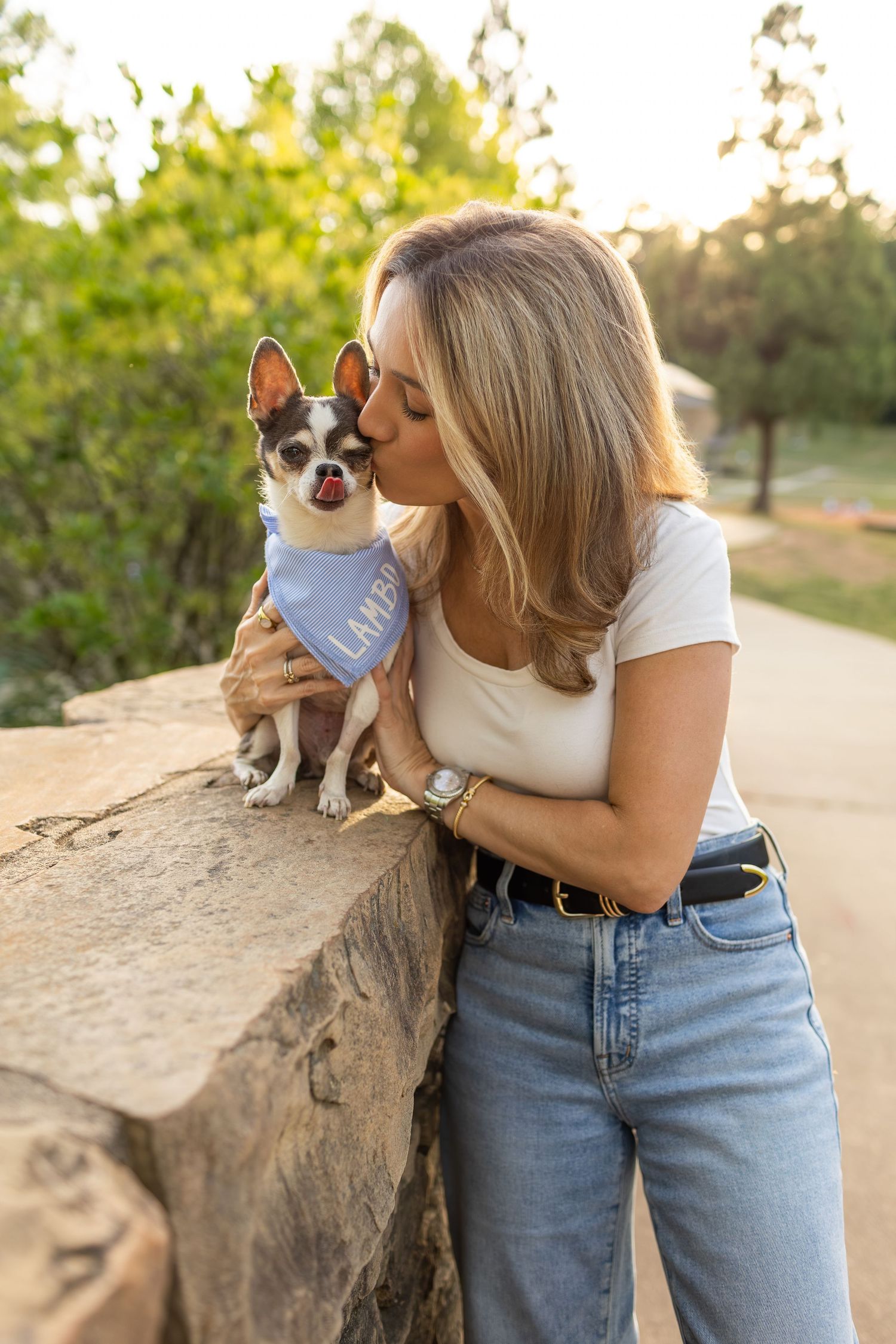 Woman in white top and jeans kissing a small Chihuahua wearing a blue bandana on a stone wall outdoors.