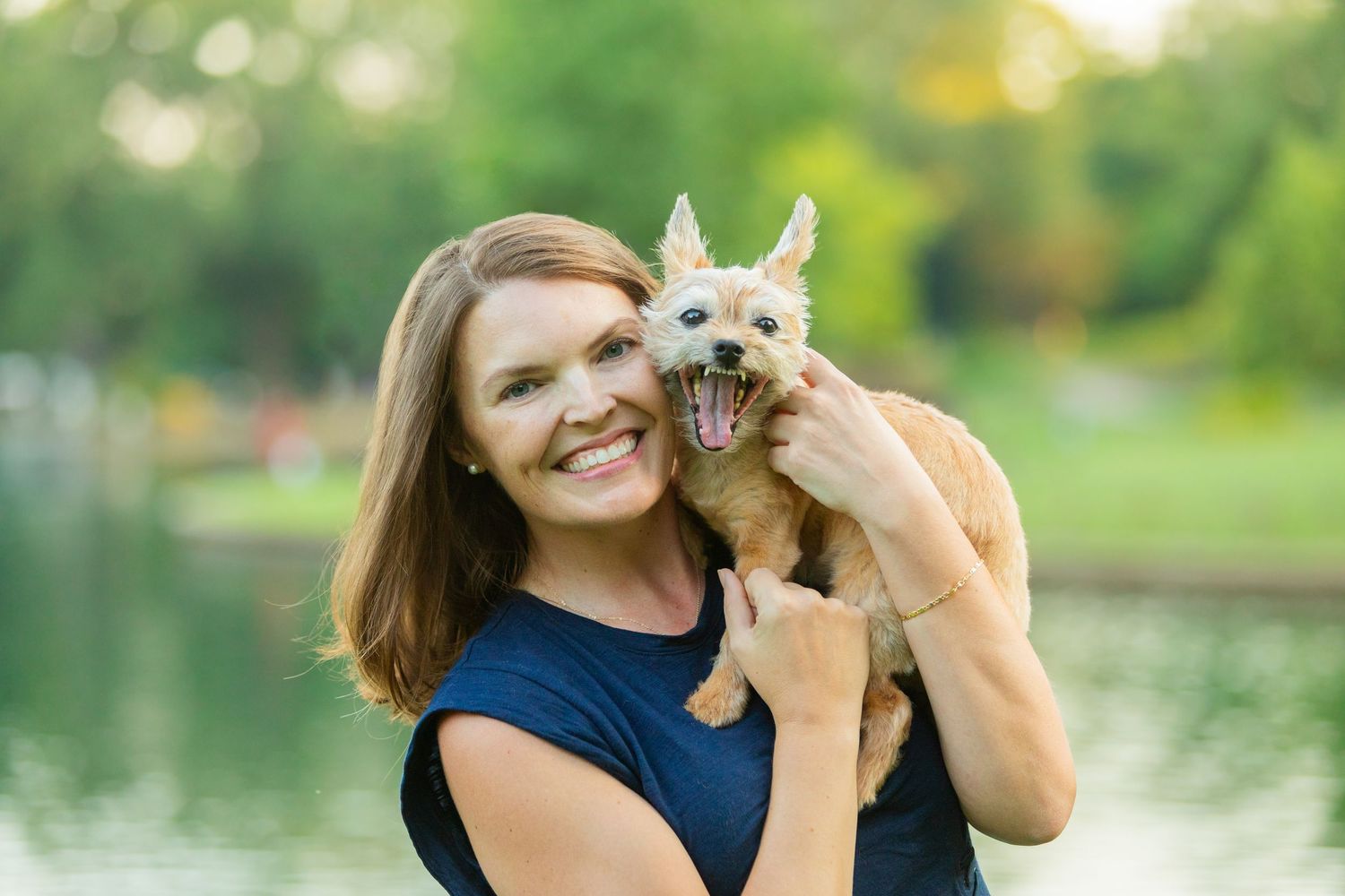 A smiling woman holds a small tan dog outdoors near a pond with lush green trees in the background.