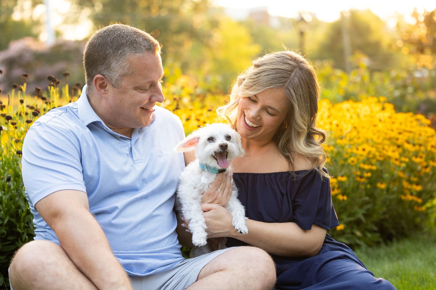 Couple sitting outdoors with small white dog amid yellow flowers in warm sunlight.