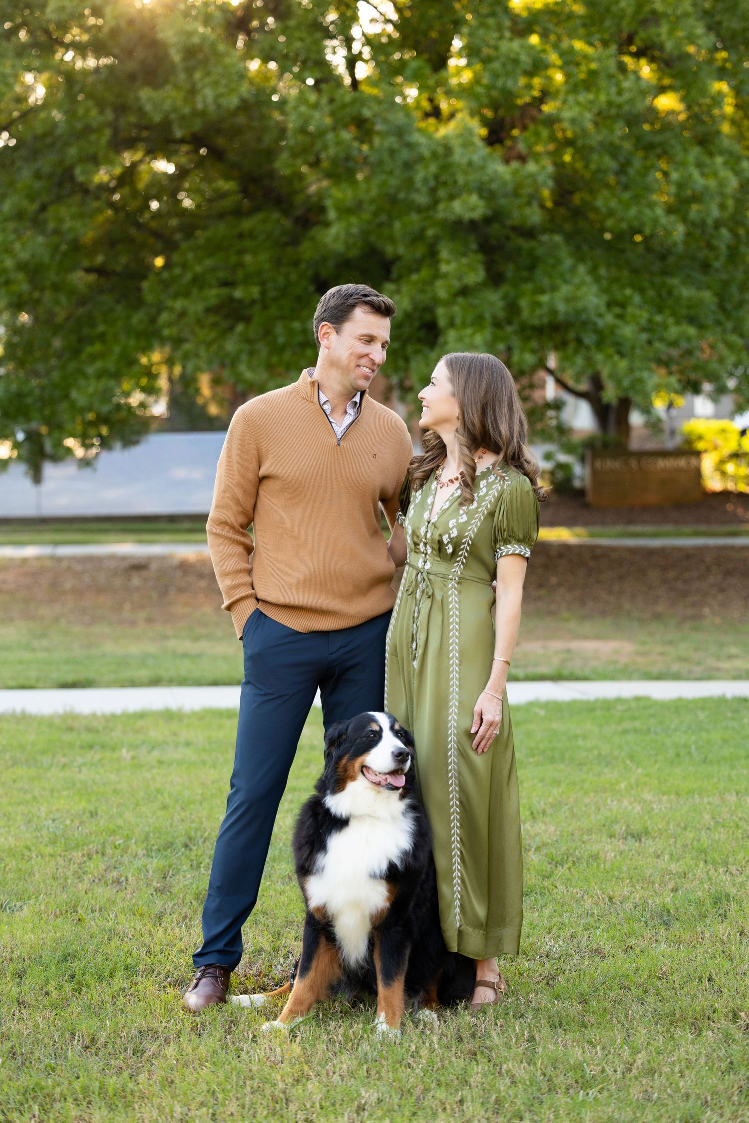 Smiling couple standing close on a sunny lawn with a Bernese Mountain Dog at their feet, surrounded by large green trees.