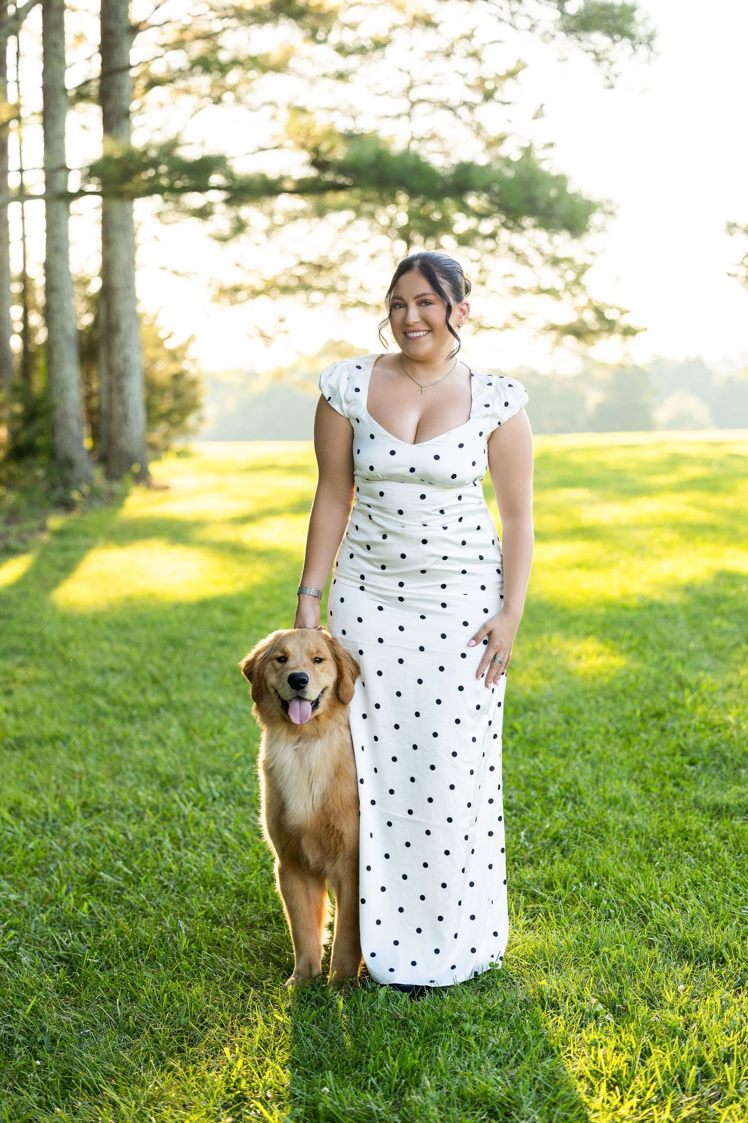 Woman in white polka dot dress smiling outdoors with golden retriever on green grass near trees.