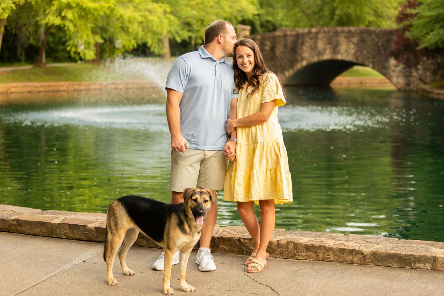 A couple sits together with their German Shepherd pet by a pond in a scenic park setting.