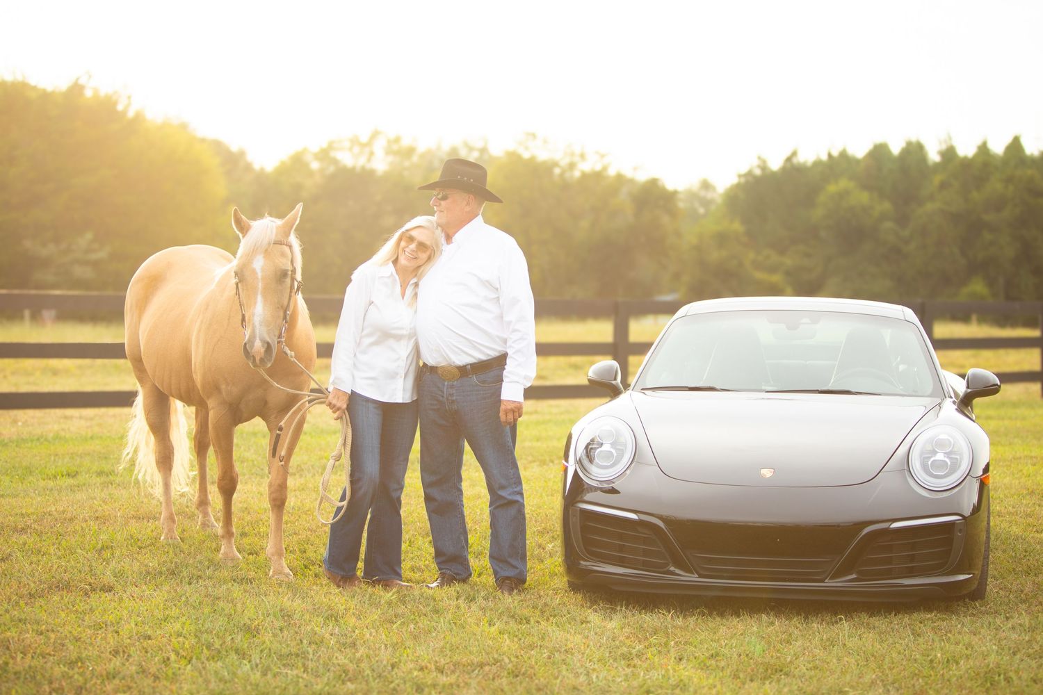 A couple stands between their horse and Porsche sports car in a sunlit field at sunset.