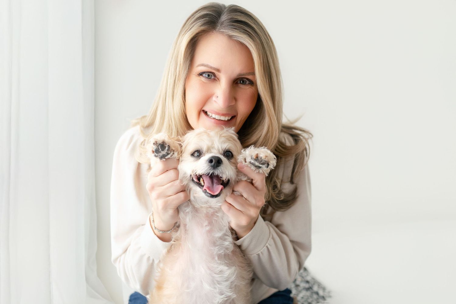 Smiling blonde woman holding up her small fluffy dog's paws toward the camera in a bright room.