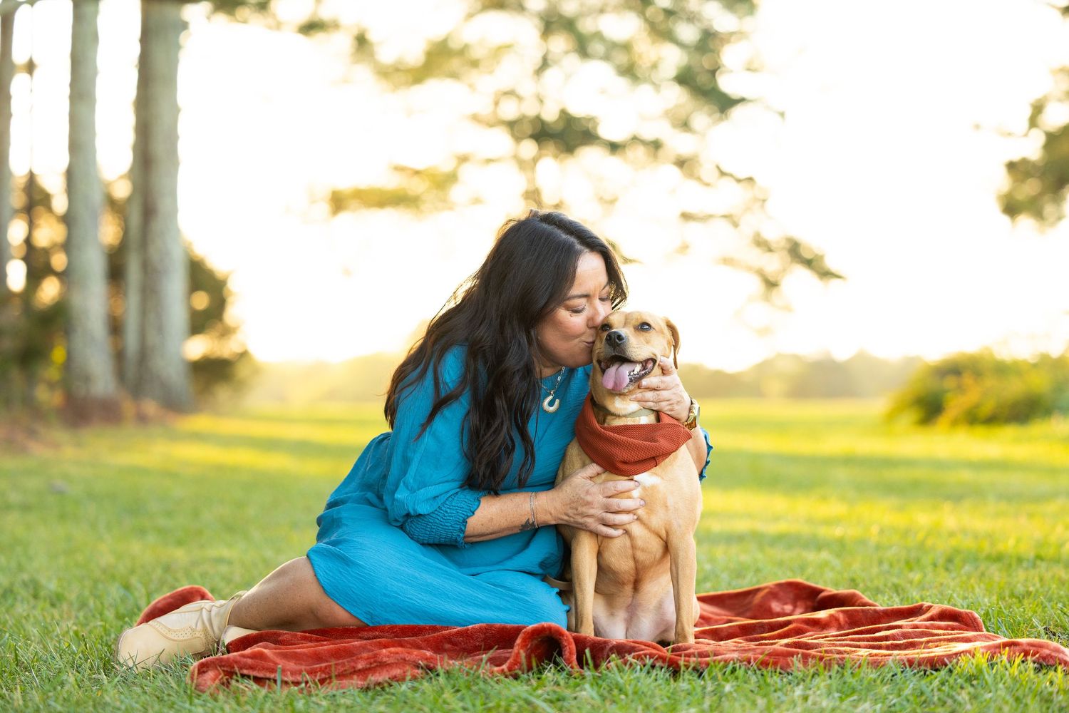 Person and golden retriever dog laying together on a red blanket in a sunny grassy field at sunset.